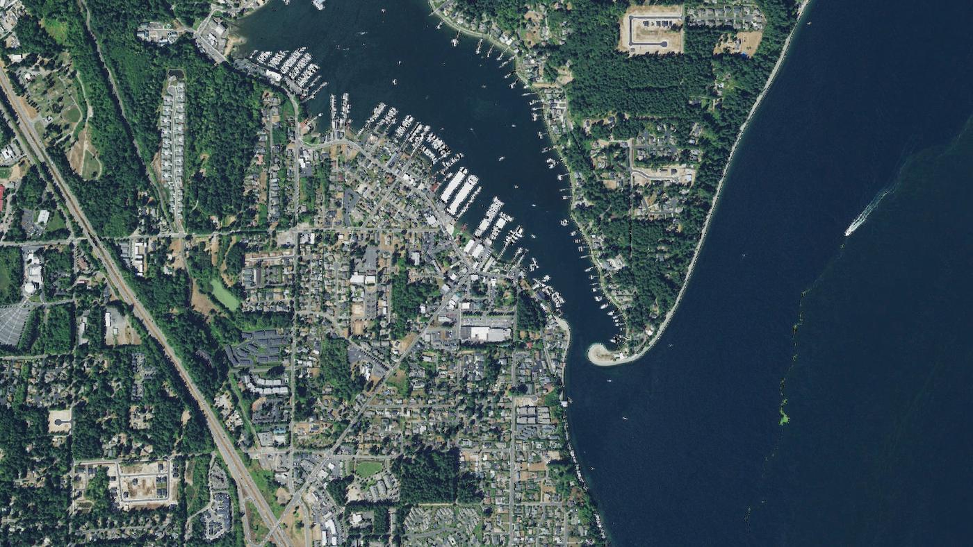 Aerial of Gig Harbor: the protected harbor lined with marinas and moored boats, downtown along the shoreline, hillside neighborhoods above, and the sandspit at the harbor mouth opening to Puget Sound