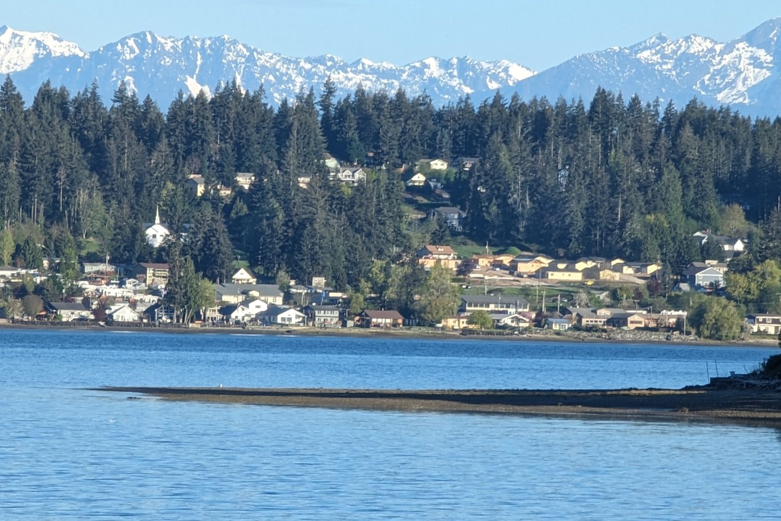 Allyn's housing stock up close: waterfront homes along North Bay, the white steeple of the historic Allyn chapel, and houses climbing the wooded hill behind SR-3, with the Olympic Mountains above