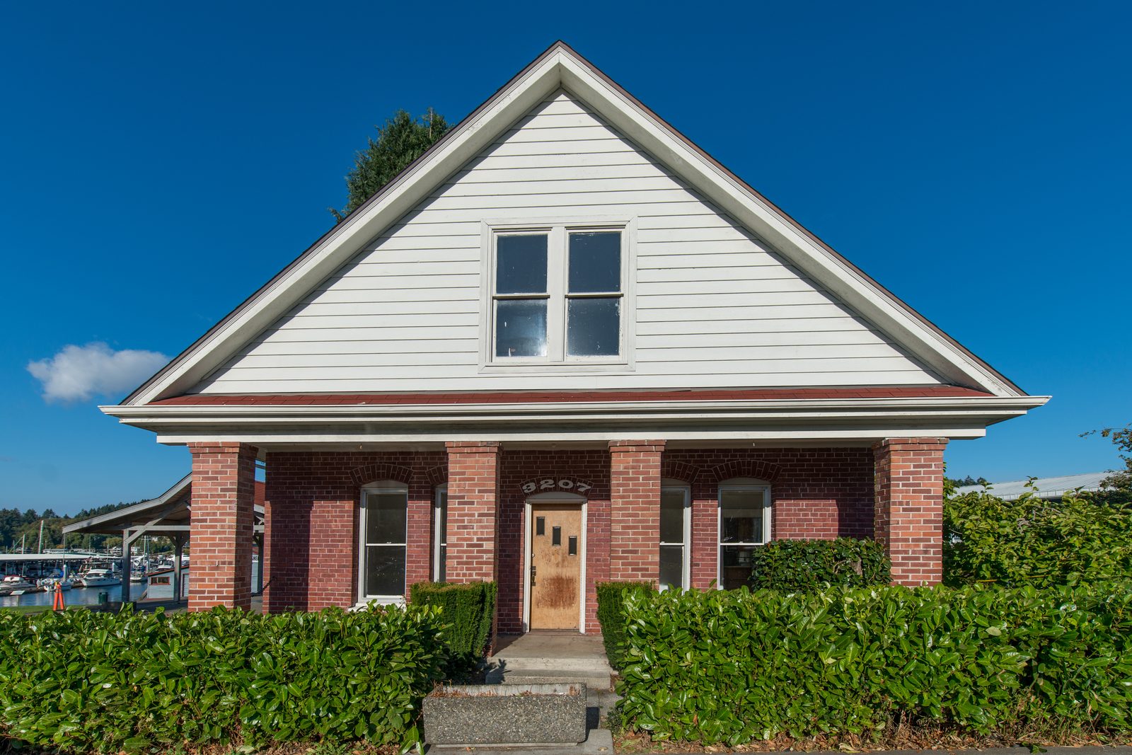 The 1908 Skansie house at Skansie Brothers Park, 3207 Harborview Drive: brick porch columns, white clapboard gable, and a laurel hedge, with the masts of the marina rising just beyond the yard