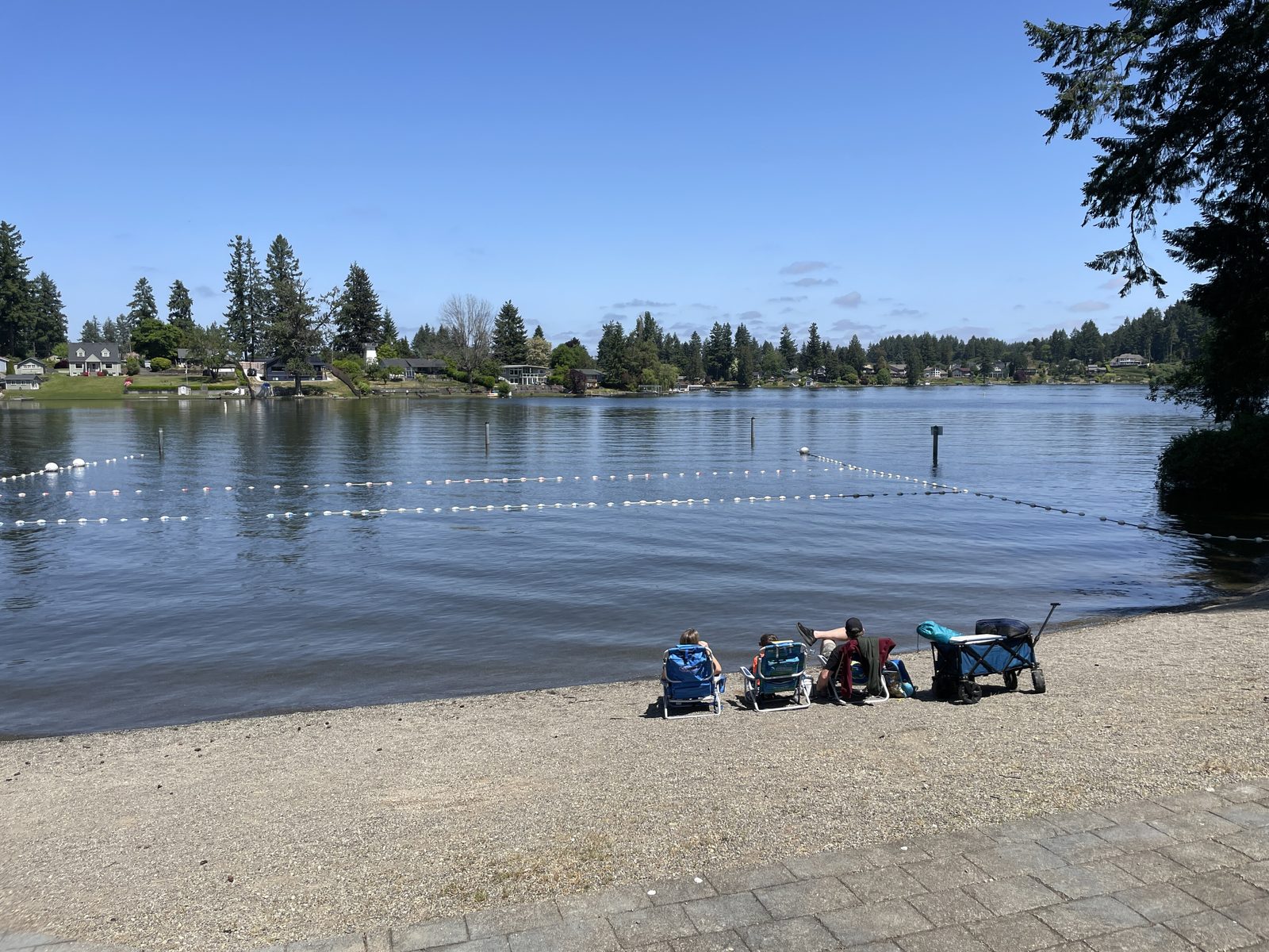 A family with camp chairs at the Long Lake Park swim beach in Lacey, lakeside homes across the water