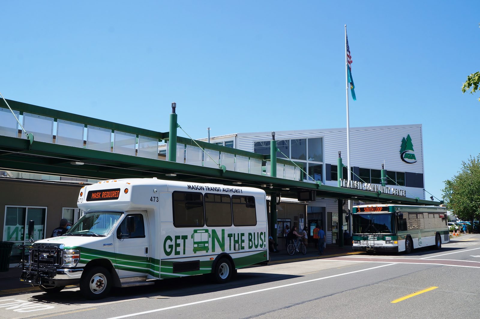 Mason Transit Authority buses at the Transit-Community Center on Railroad Avenue in downtown Shelton, with riders and a cyclist on the sidewalk