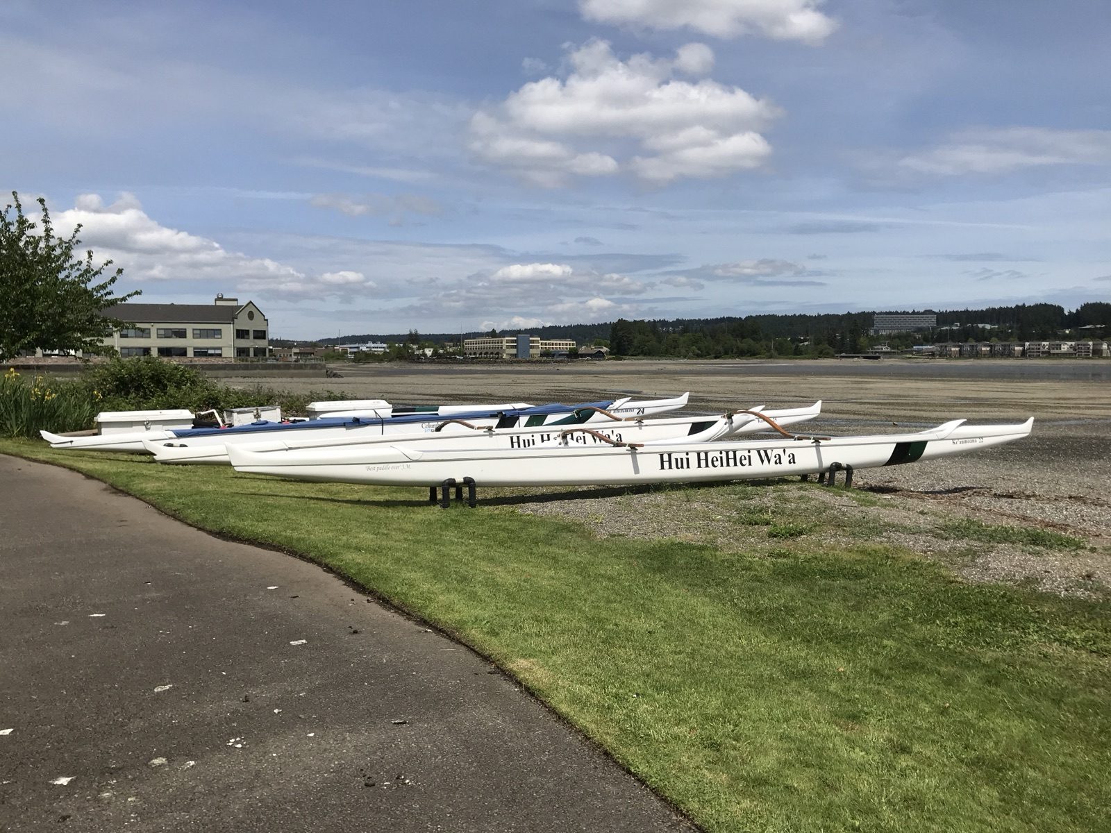 Outrigger canoes beside a waterfront path in Silverdale, with buildings across the inlet at low tide