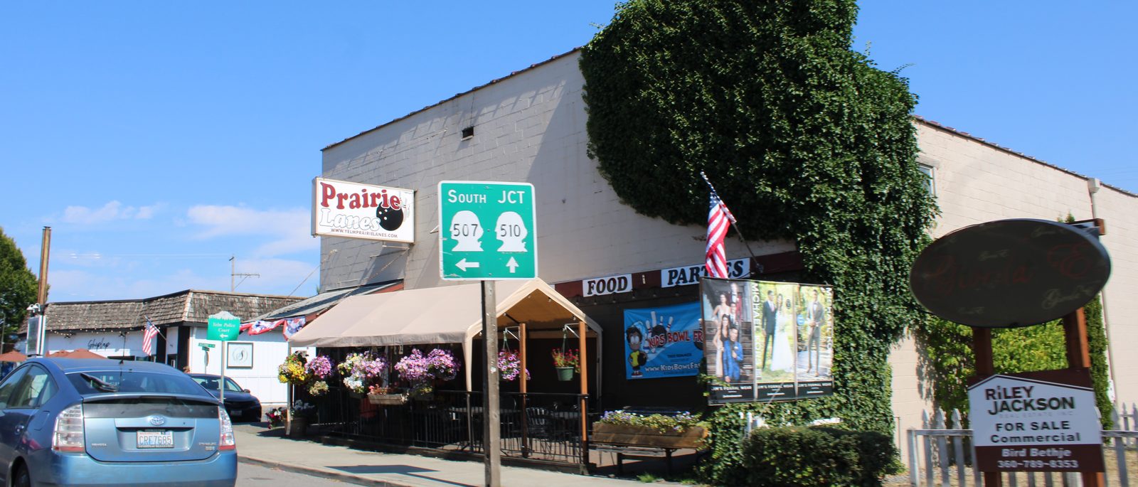 Downtown Yelm along Yelm Avenue: the Prairie Lanes block with sidewalk flower baskets, patio seating, and the flag out front
